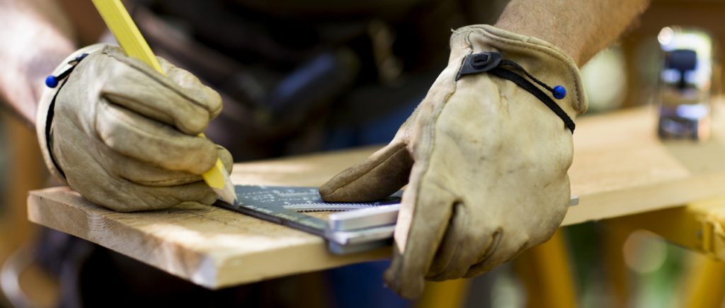 Cropped view of a carpenter marking a measurement on a wooden plank. Horizontal shot.